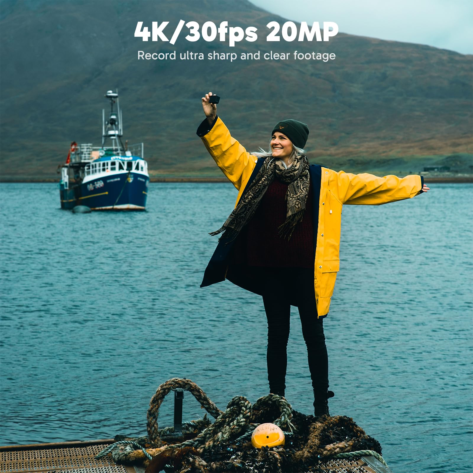 Une femme en veste jaune sur un dock en bois tient un petit appareil photo, avec un bateau et des montagnes.