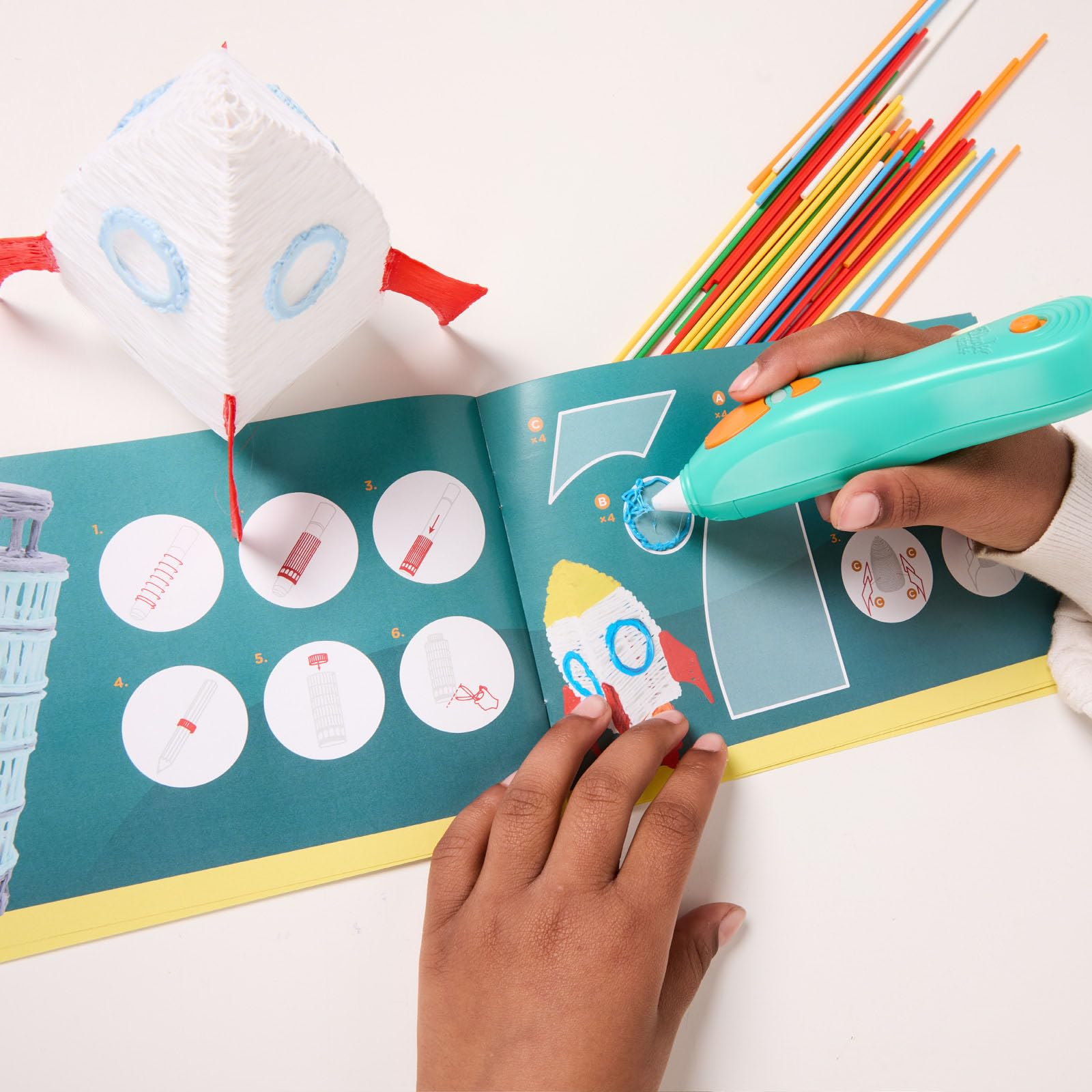 Un enfant utilise un stylo 3D pour dessiner une fusée d'après un livret, avec des tiges colorées.
