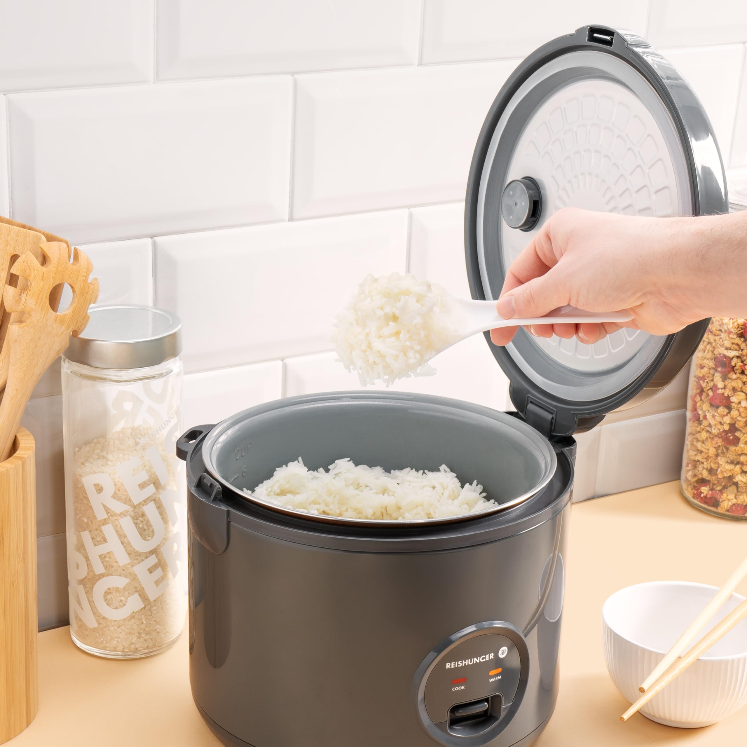Ein Reiskocher in einer Küche, in der Hände Reis mit einem Löffel schöpfen.