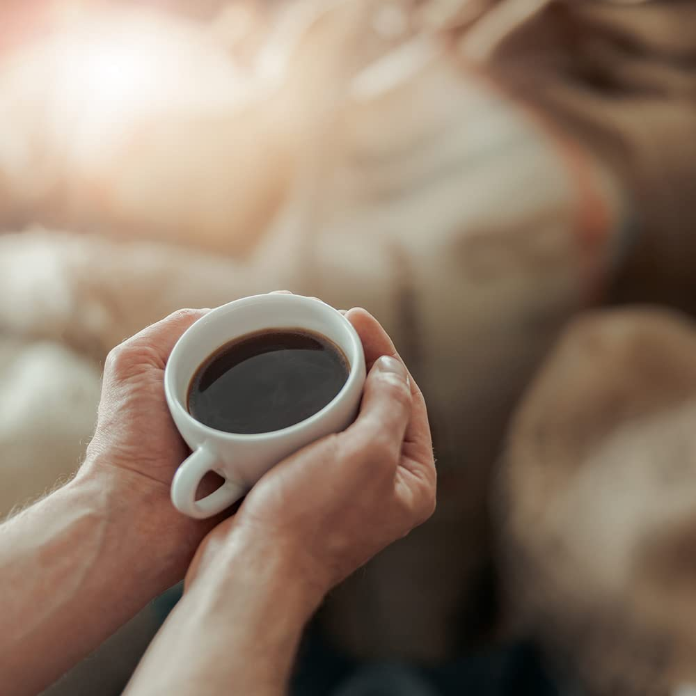 Mani che tengono una tazza bianca di caffè nero su uno sfondo caldo e sfocato.