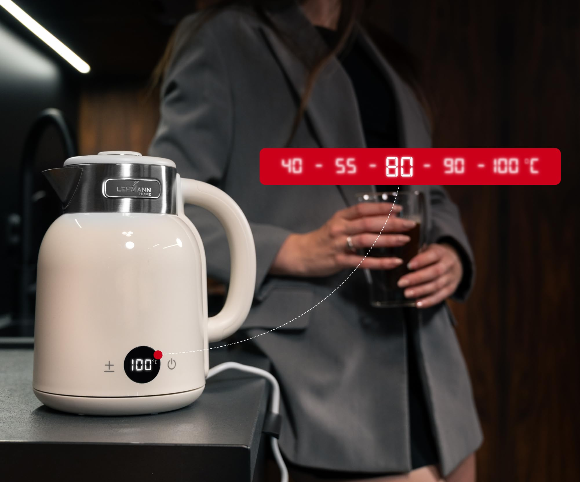 Un bollitore elettrico bianco su un banco con display digitale, persona con bicchiere sullo sfondo.