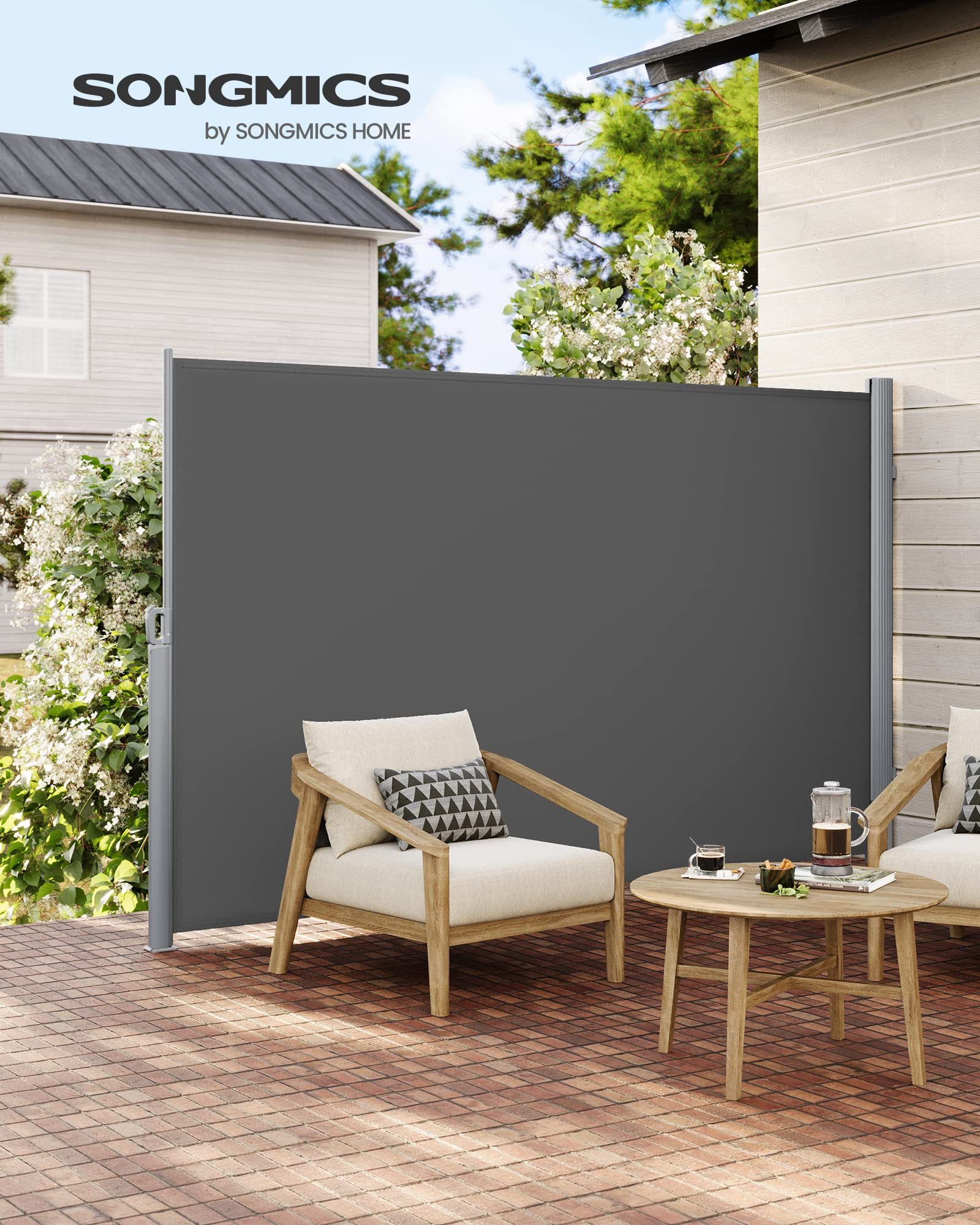 Eine graue Seitenmarkise auf einer Terrasse mit Holzstühlen, einem runden Tisch und blühenden Büschen an einer Hauswand.