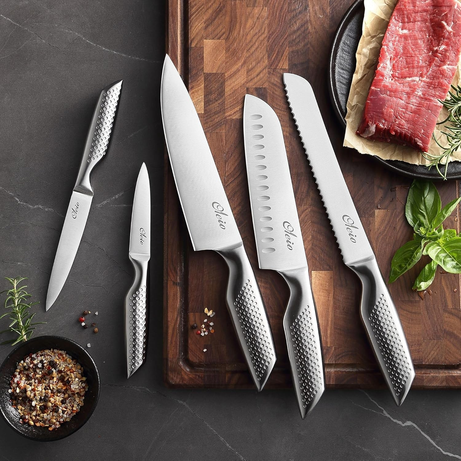 Fünf Edelstahlmesser mit strukturierten Griffen auf einem Holzbrett neben rohem Fleisch, Kräutern und Gewürzen.