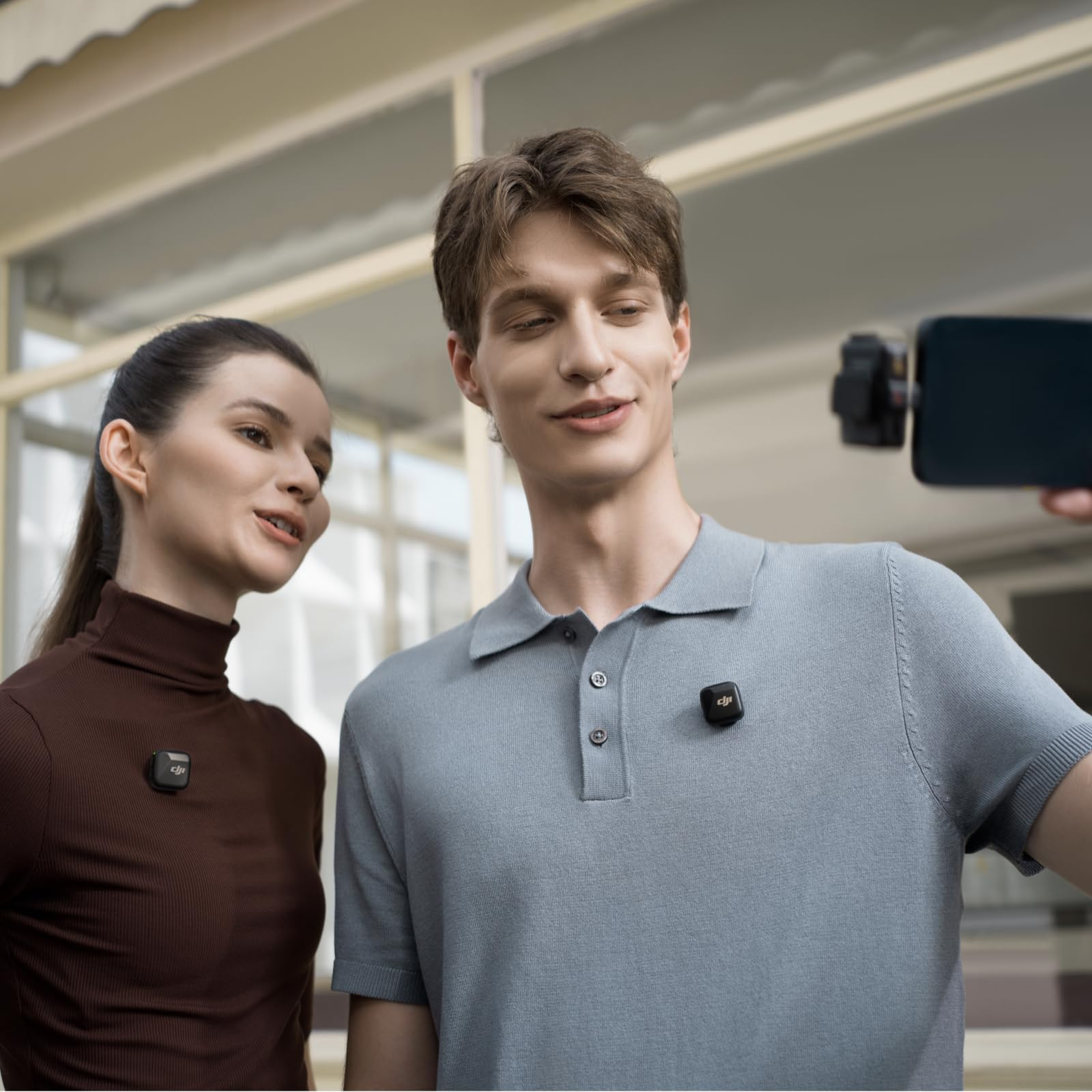 Un homme et une femme portant de petits micros-cravates DJI prennent un selfie avec un smartphone à l'extérieur.