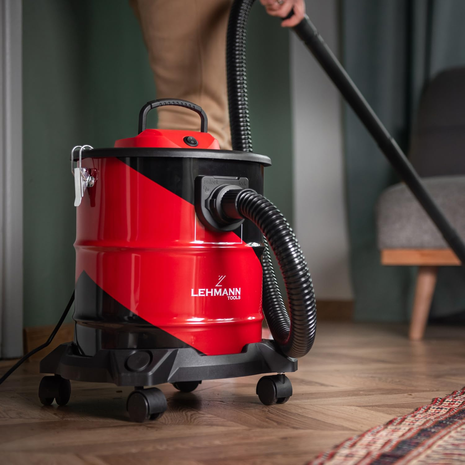 Un aspirapolvere/liquidi Lehmann Tools rosso e nero su pavimento in legno, con una persona che aspira sullo sfondo.
