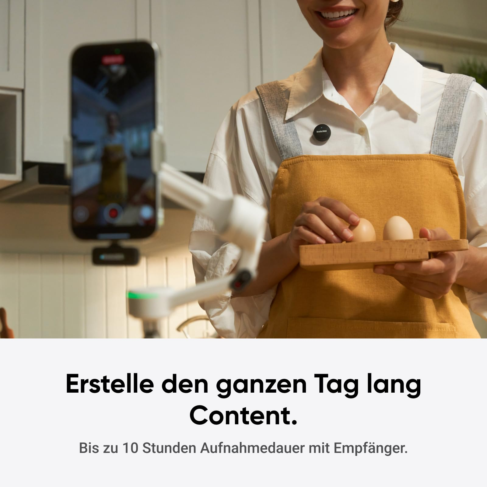 Eine Frau mit gelber Schürze hält Eier, während ein Smartphone auf einem Stativ sie in einer Küche filmt.