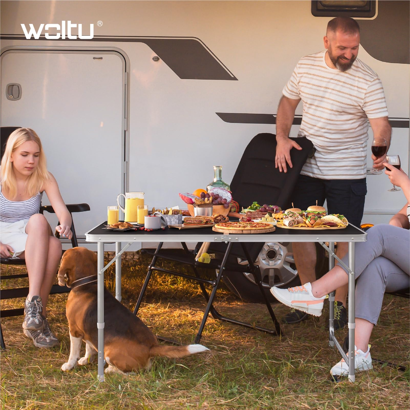 Eine Familie und ein Beaglohund genießen ein Picknick mit Essen und Wein an einem Wohnmobil.