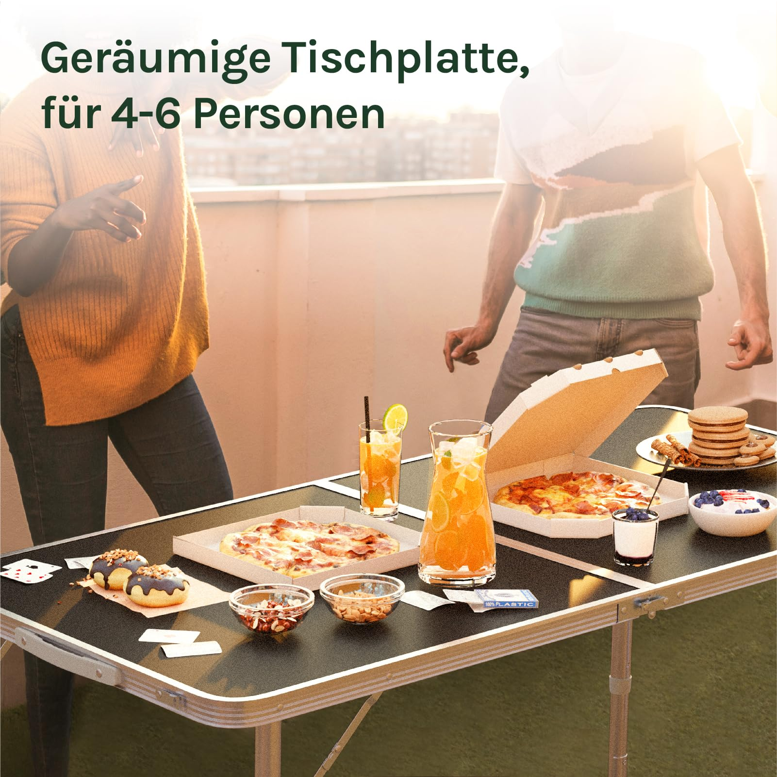 Ein Tisch mit Pizza, Getränken und Snacks steht auf einem Balkon, während zwei Personen im Hintergrund stehen.