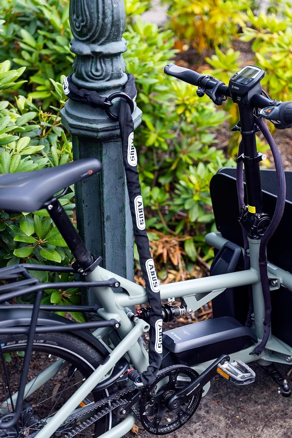 Un vélo électrique gris attaché avec une chaîne ABUS noire à un poteau vert.