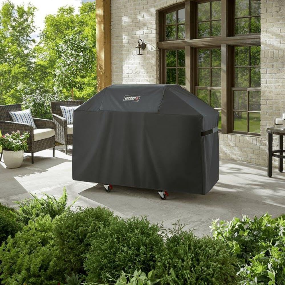 Ein abgedeckter Weber-Grill steht auf einer Terrasse mit Korbstühlen und üppig grünen Pflanzen im Vordergrund.