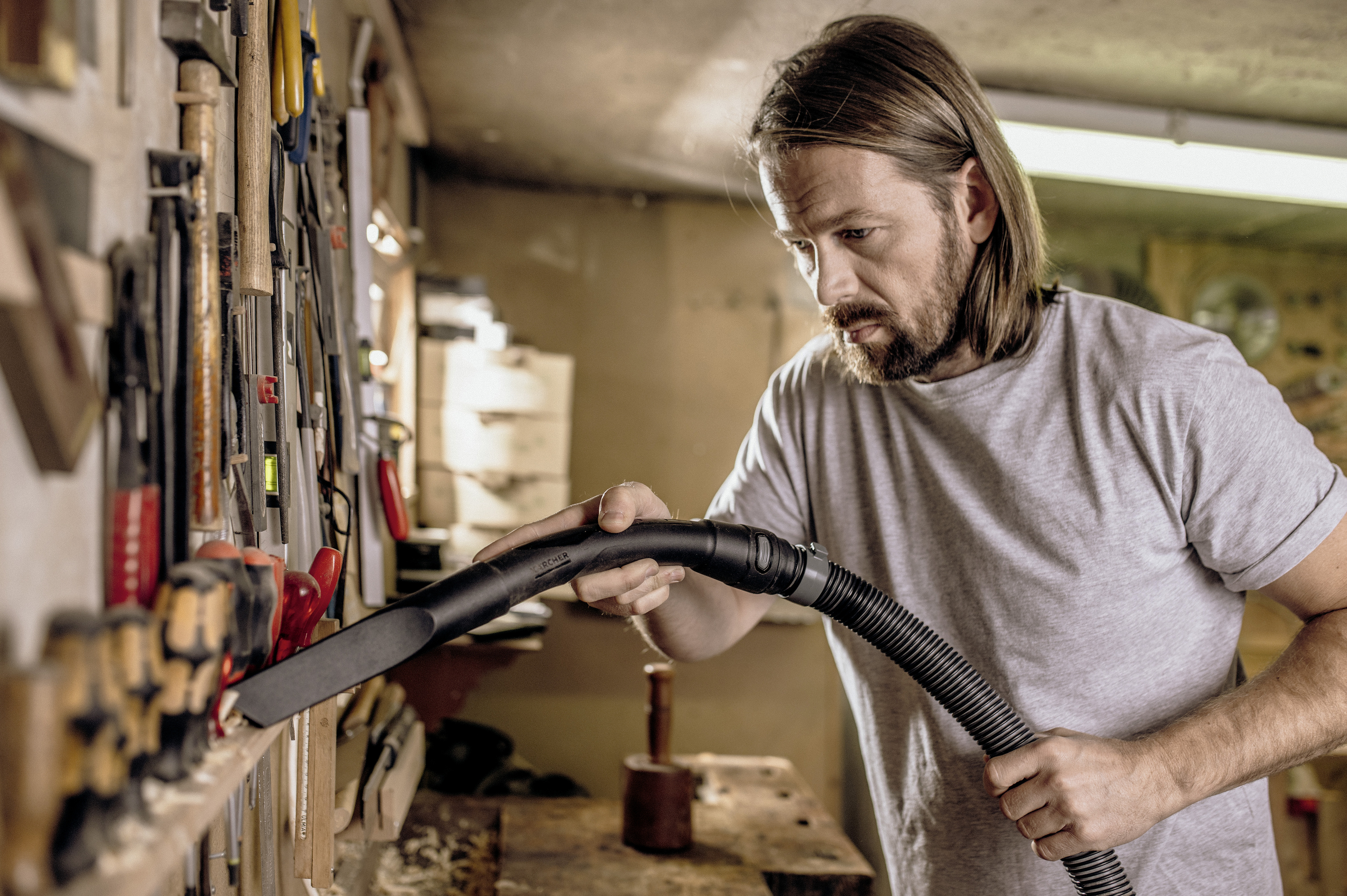 Ein Mann mit langen Haaren und Bart reinigt mit einem Staubsaugerschlauch eine Werkzeugwand in seiner Werkstatt.