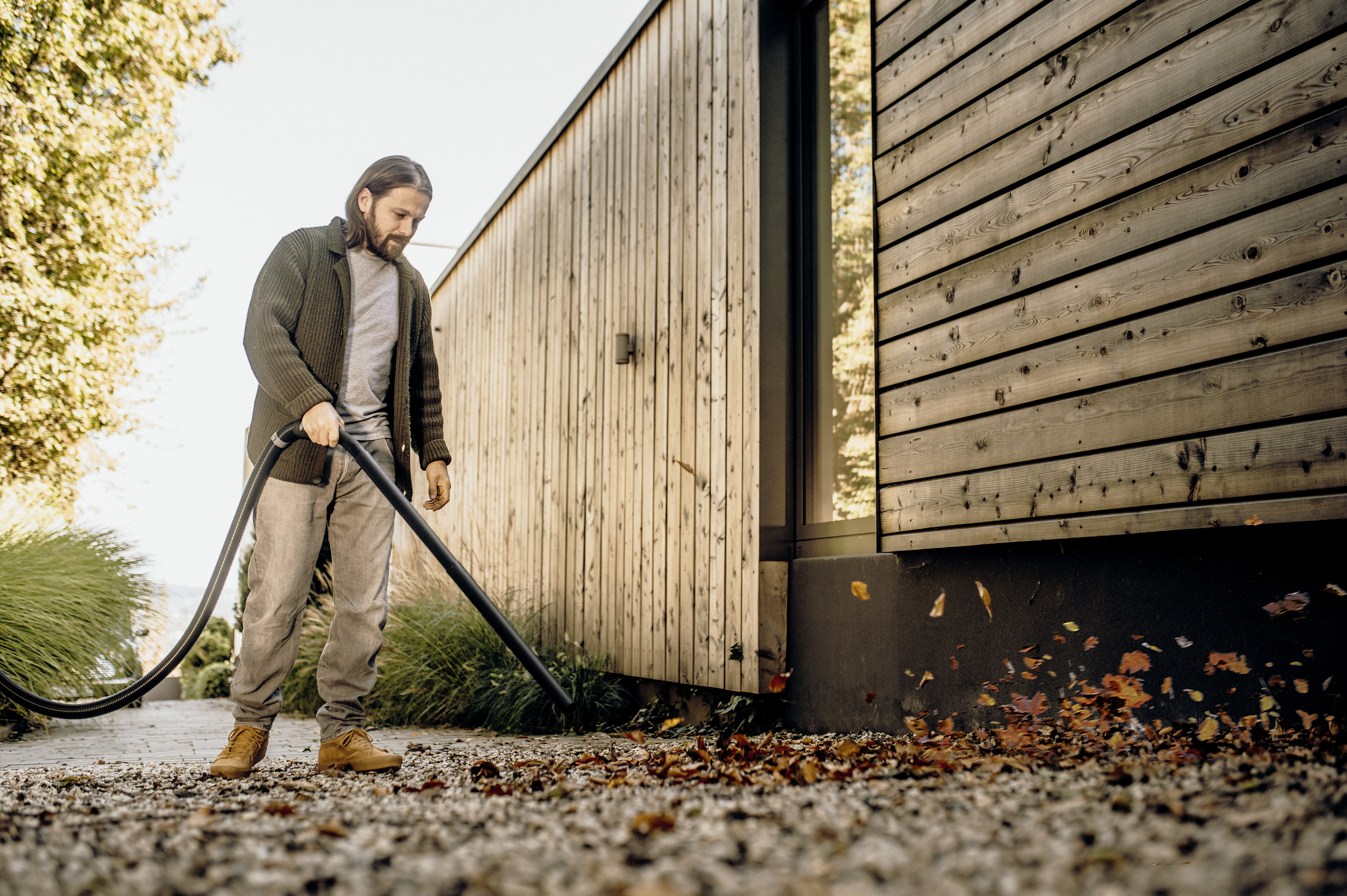 Ein Mann mit Bart und Strickjacke entfernt herbstliche Blätter mit einem Bläser vor einem modernen Holzhaus.