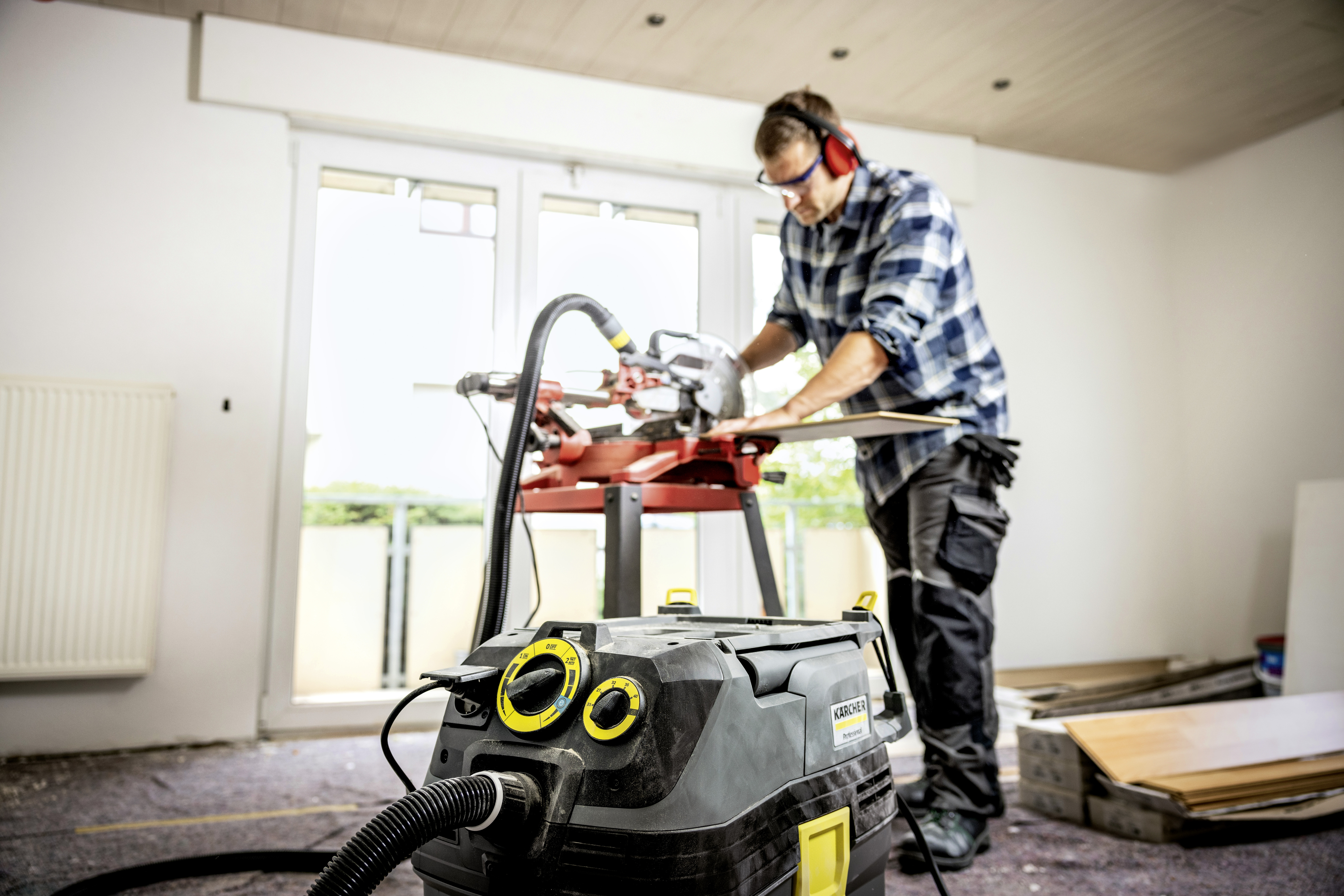 Ein Handwerker sägt Holz in einem Raum mit einem großen Staubsauger im Vordergrund.