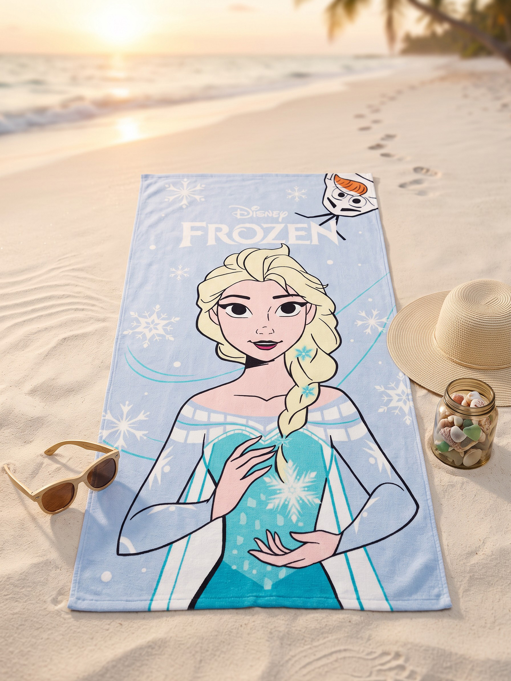 Ein Elsa-Strandtuch liegt im Sand neben einer Sonnenbrille, einem Hut und einem Glas mit Muscheln.