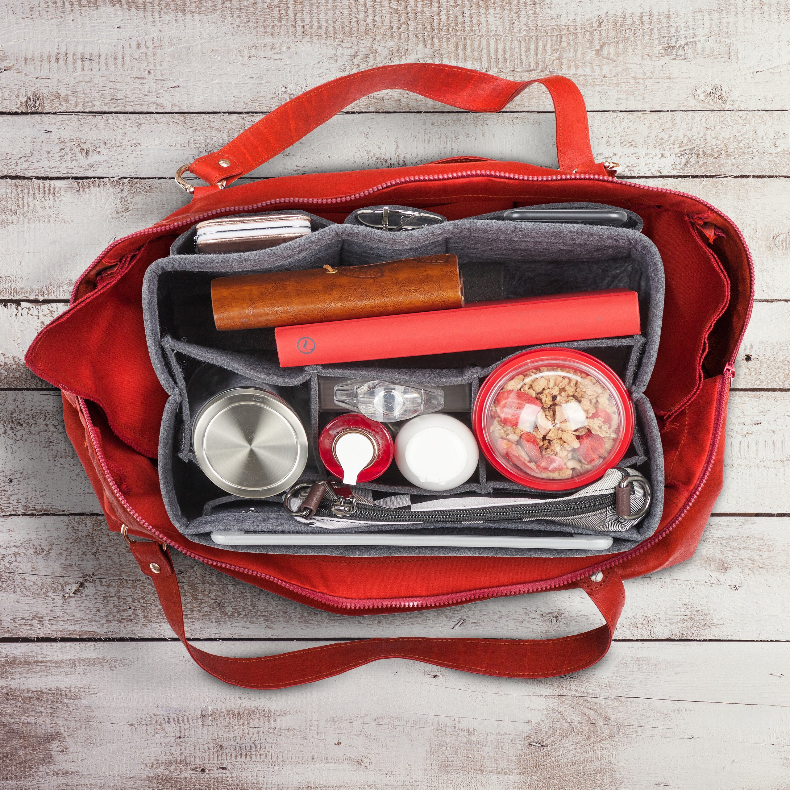 Eine rote Tasche von oben, mit grauem Filzorganizer gefüllt mit Büchern, Flaschen und Snacks auf Holz.