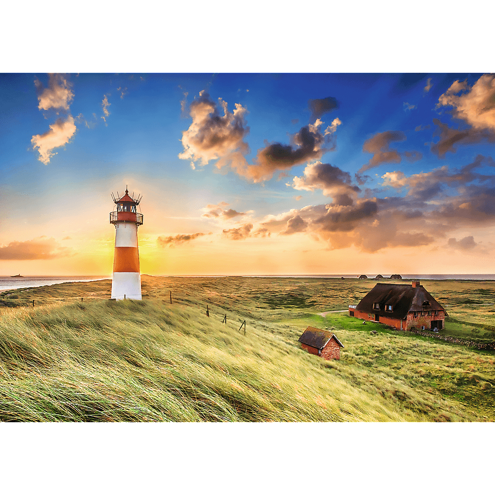 Ein rot-weißer Leuchtturm steht bei Sonnenuntergang auf einer Düne neben roten Backsteinhäusern.