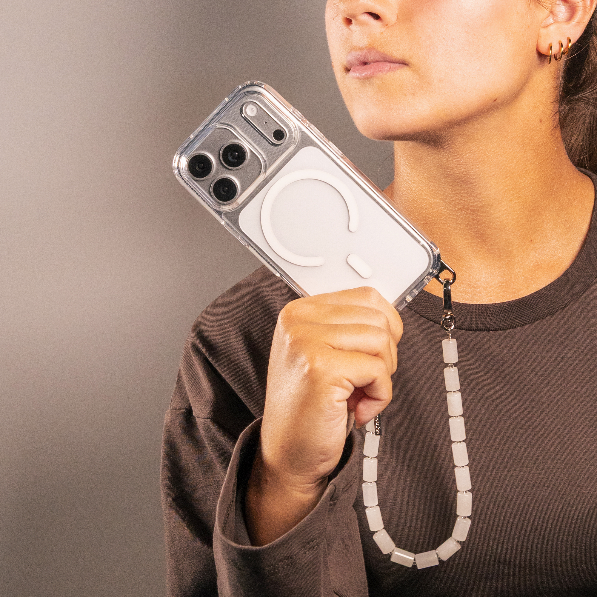A person holding a smartphone with a clear case and beaded strap against a solid grey background.