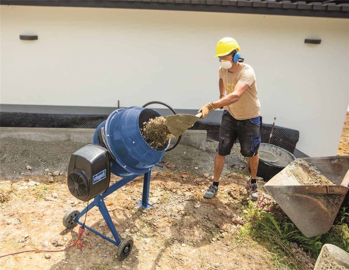 Un lavoratore con elmetto giallo e mascherina usa una pala per svuotare una betoniera blu.