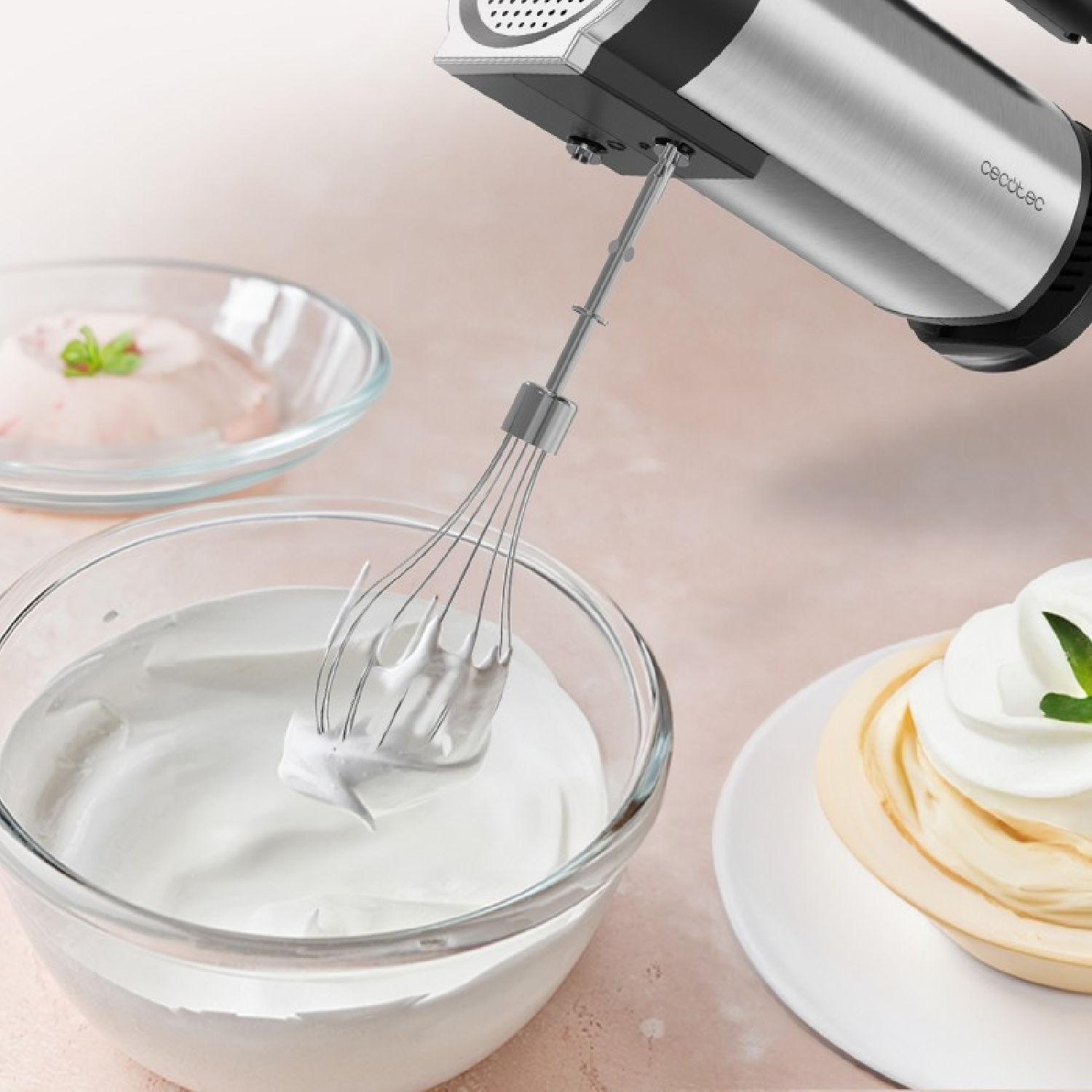 Una batidora de mano bate nata blanca en un bol de cristal, con un postre y un pastel cerca.