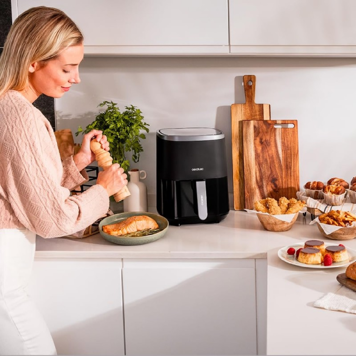 Una mujer de rosa sazona salmón en una cocina luminosa junto a una freidora de aire y alimentos.