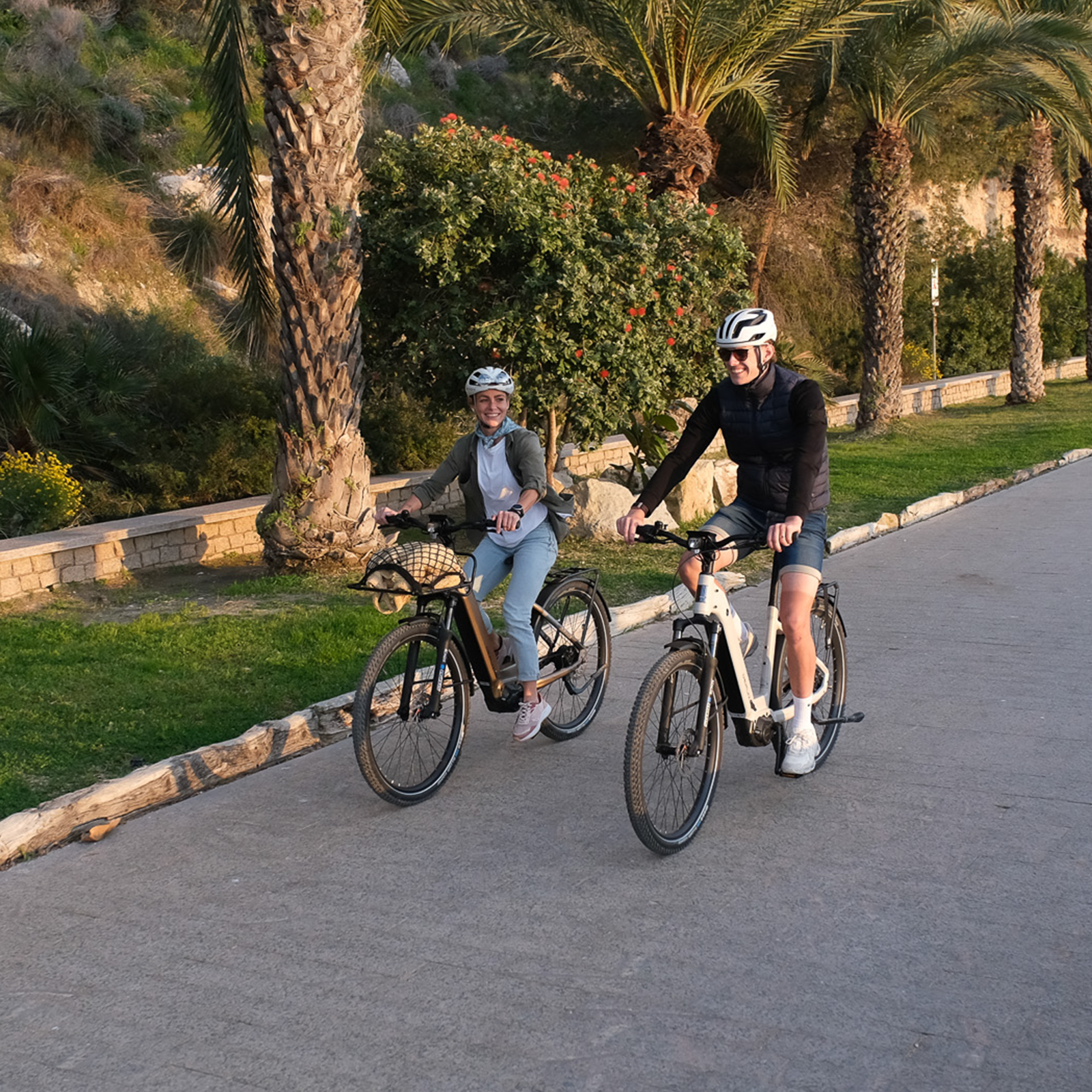 Ein Paar fährt bei Sonnenuntergang mit E-Bikes auf einem Weg neben Palmen und Blumen.