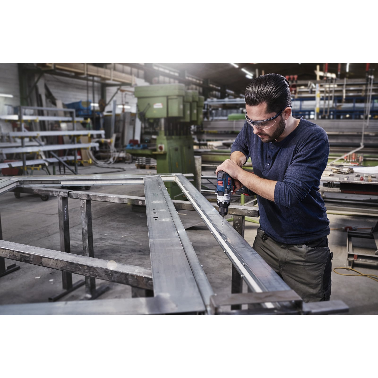 Ein Arbeiter mit Schutzbrille bohrt in einer Fabrik mit einer blauen Bohrmaschine in ein Metallteil.