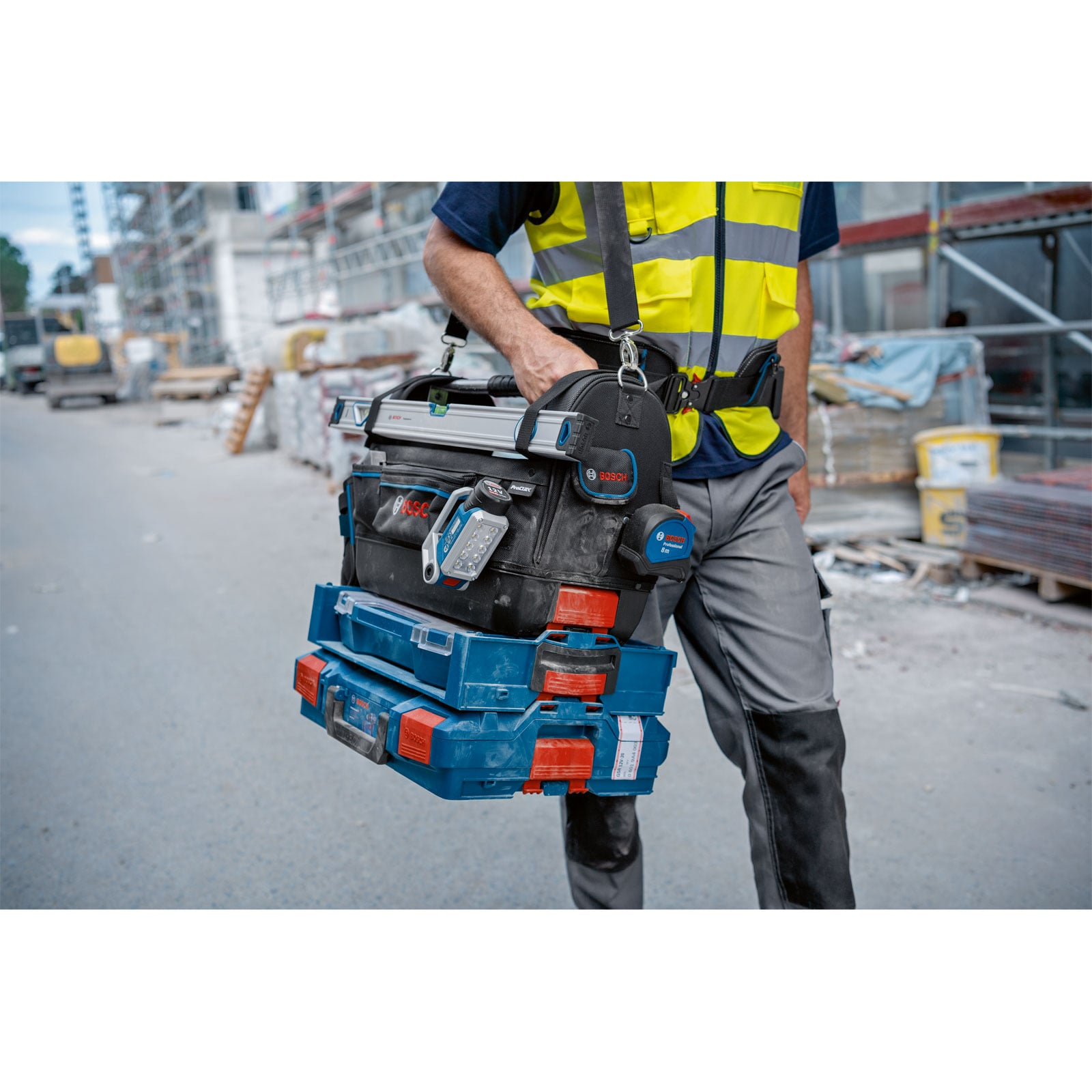 Ein Arbeiter mit Warnweste trägt eine Bosch-Werkzeugtasche mit gestapelten Koffern auf einer Baustelle.