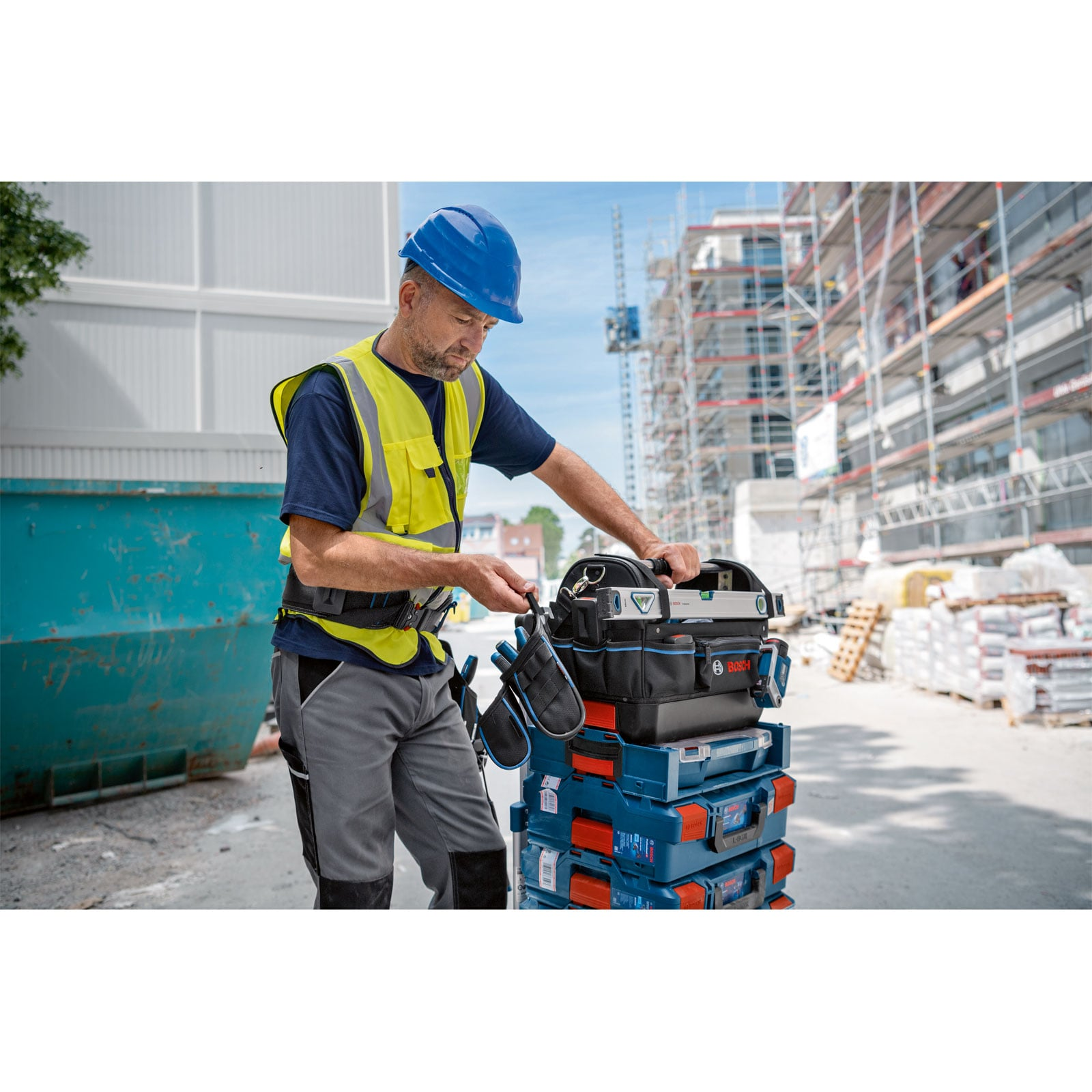 Ein Arbeiter in blauem Helm und Warnweste steht neben gestapelten Werkzeugkoffern auf einer Baustelle.