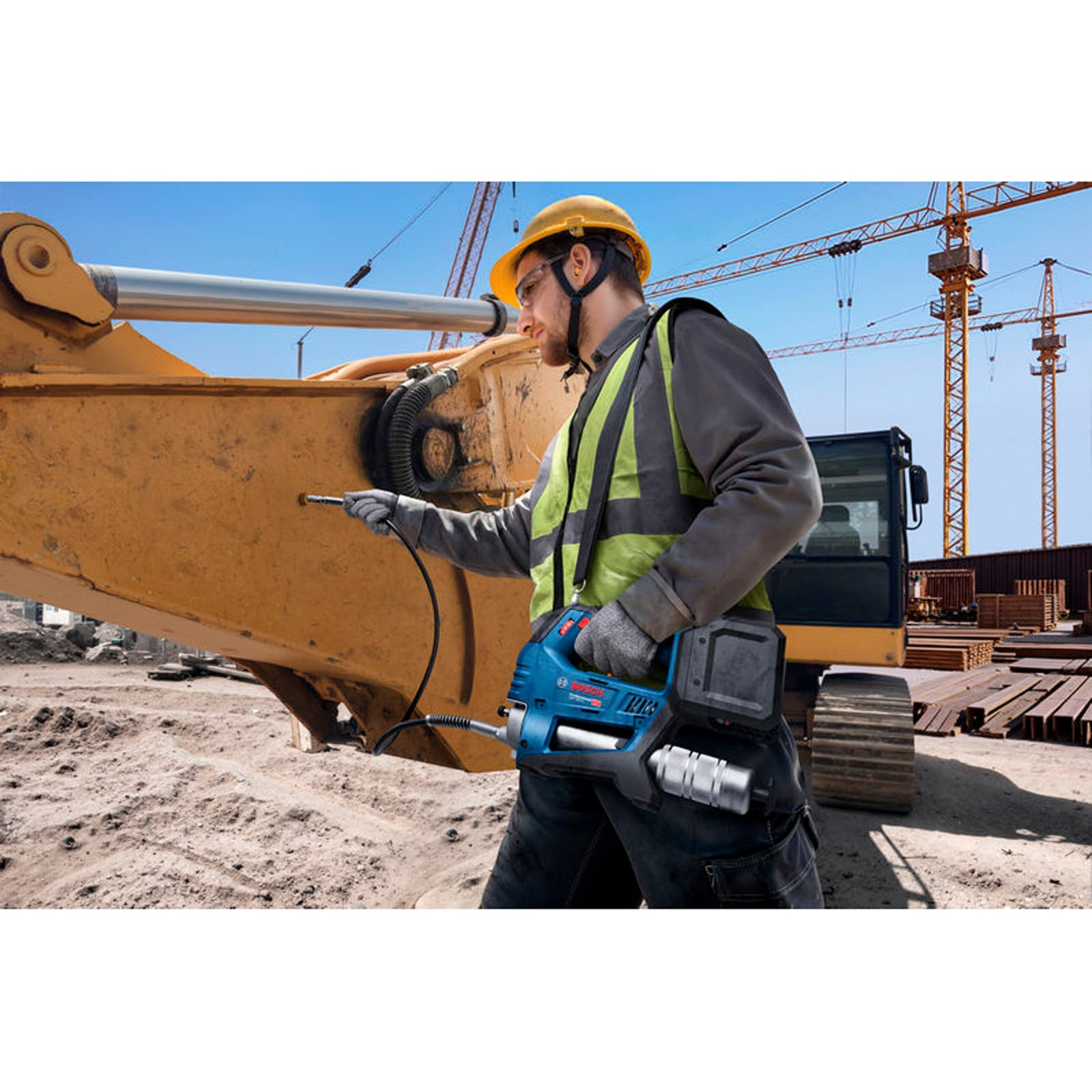 Ein Arbeiter mit gelbem Helm fettet auf einer Baustelle mit einem blauen Werkzeug einen Bagger ein.