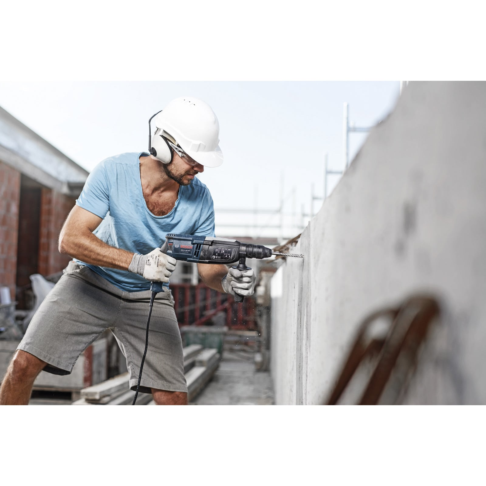 Ein Arbeiter mit weißem Schutzhelm und Schutzbrille bohrt mit einem Bohrhammer in eine Betonwand.