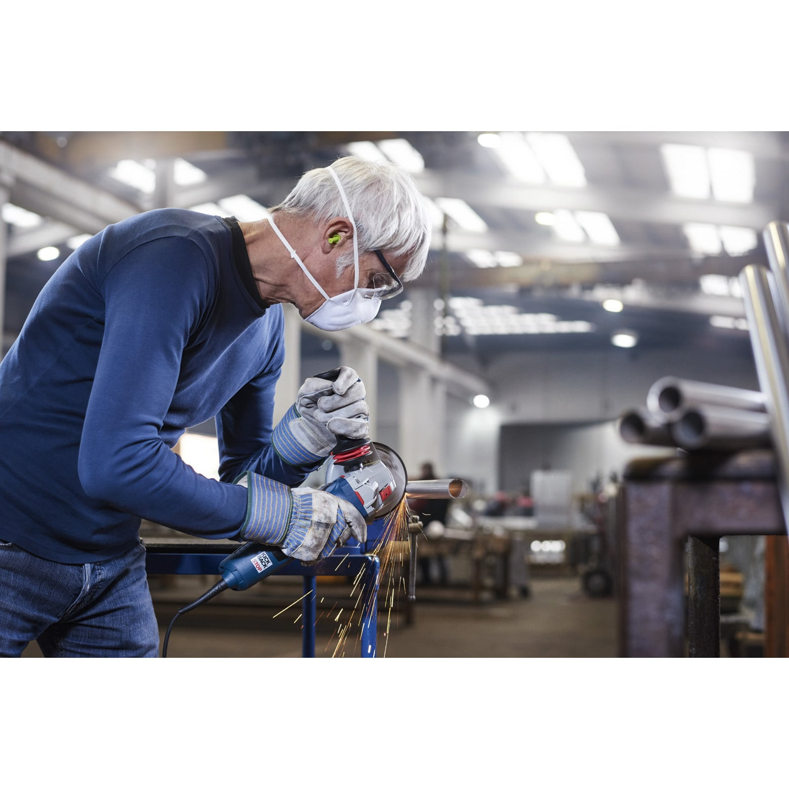 Ein älterer Handwerker schneidet mit einer Schleifmaschine ein Metallrohr, während er Schutzkleidung trägt.