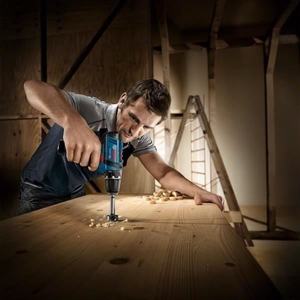 Ein Handwerker bohrt mit einer blauen Bohrmaschine ein Loch in eine helle Holztplatte in einer Werkstatt.