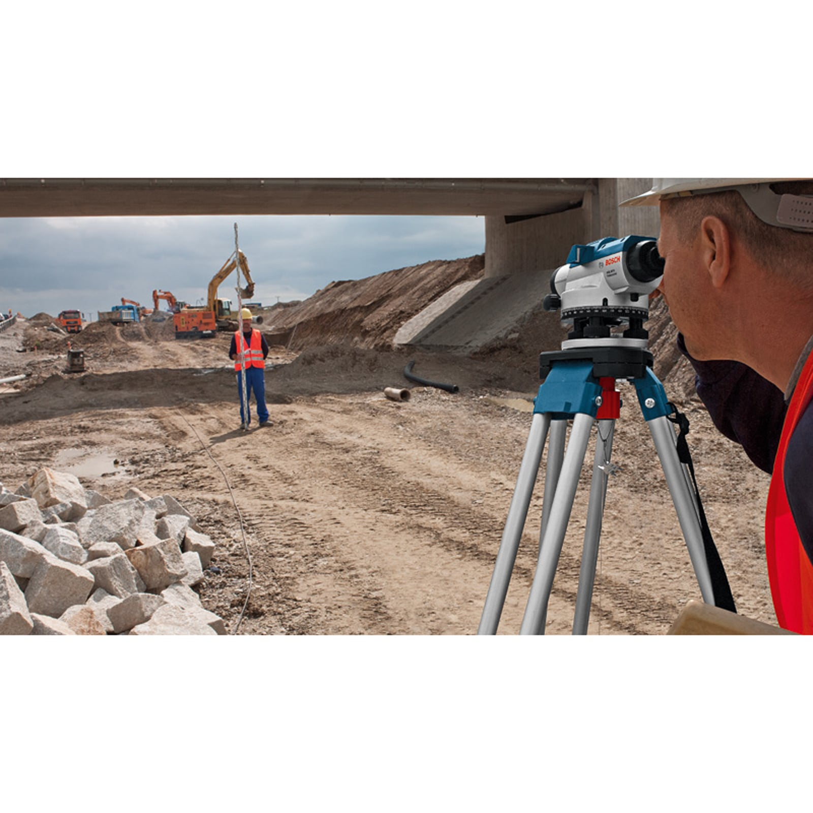 Bauarbeiter nutzt Nivelliergerät auf Baustelle, im Hintergrund weiterer Arbeiter mit Messlatte und Baufahrzeuge.