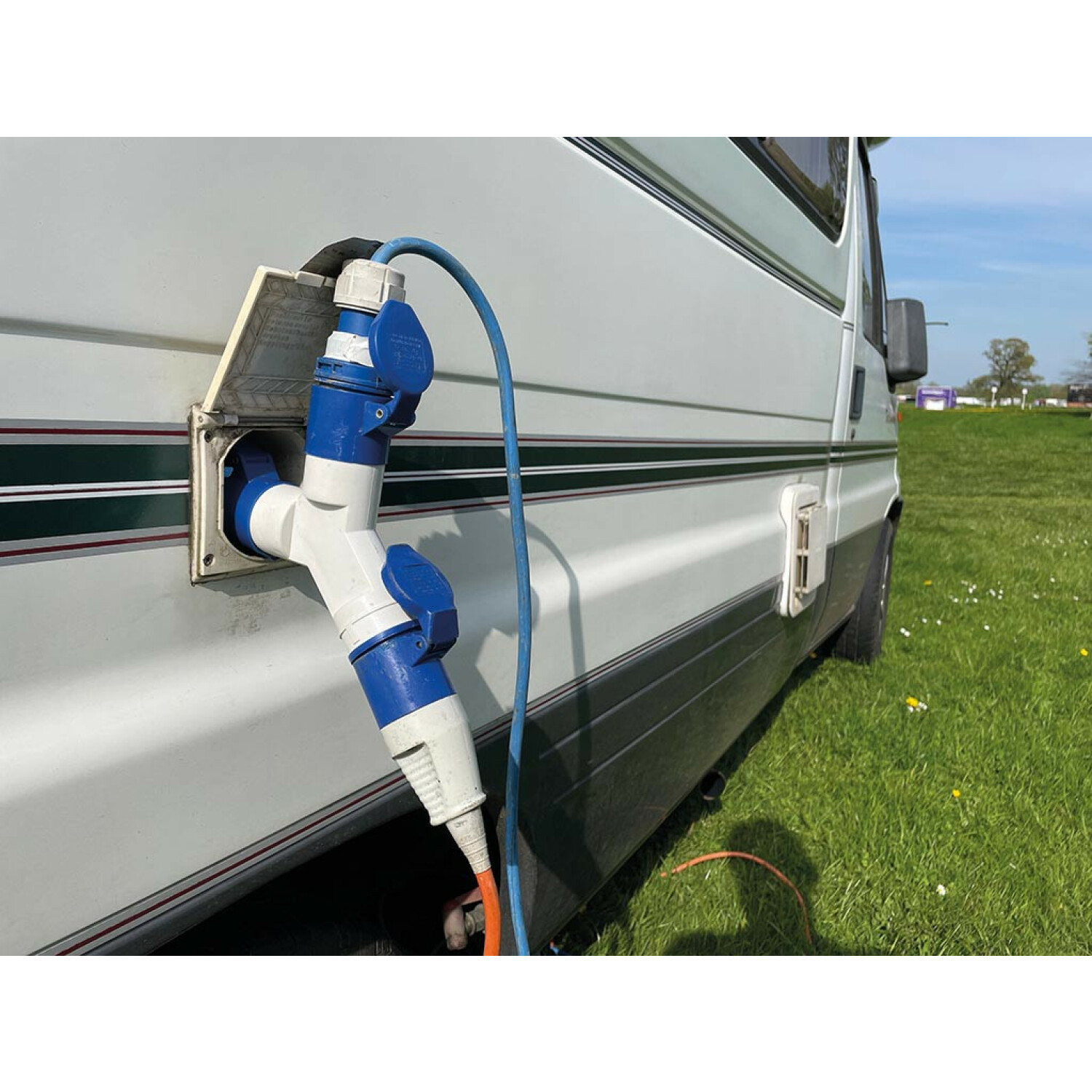 Blau-weißer Stromadapter und Kabel an einem weißen Wohnmobil auf einer grünen Wiese.
