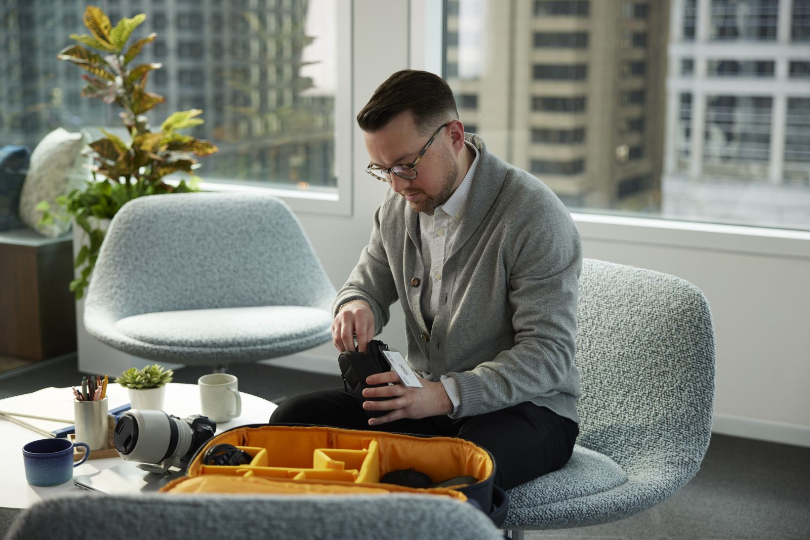 Ein Mann mit Brille und grauem Cardigan sitzt in einem Büro und packt eine Kameratasche.