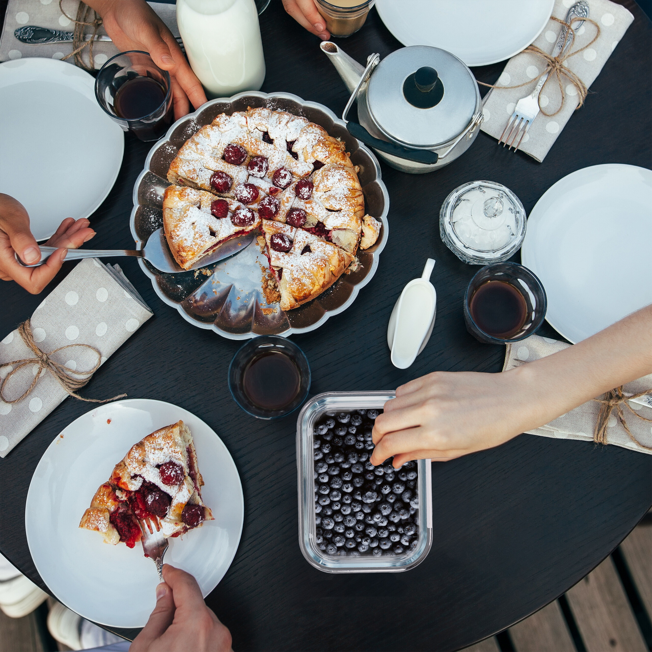 Überkopfaufnahme eines gedeckten Tisches mit Beerenkuchen, Tellern, Tassen, Teekanne und Händen, die das Essen greifen.