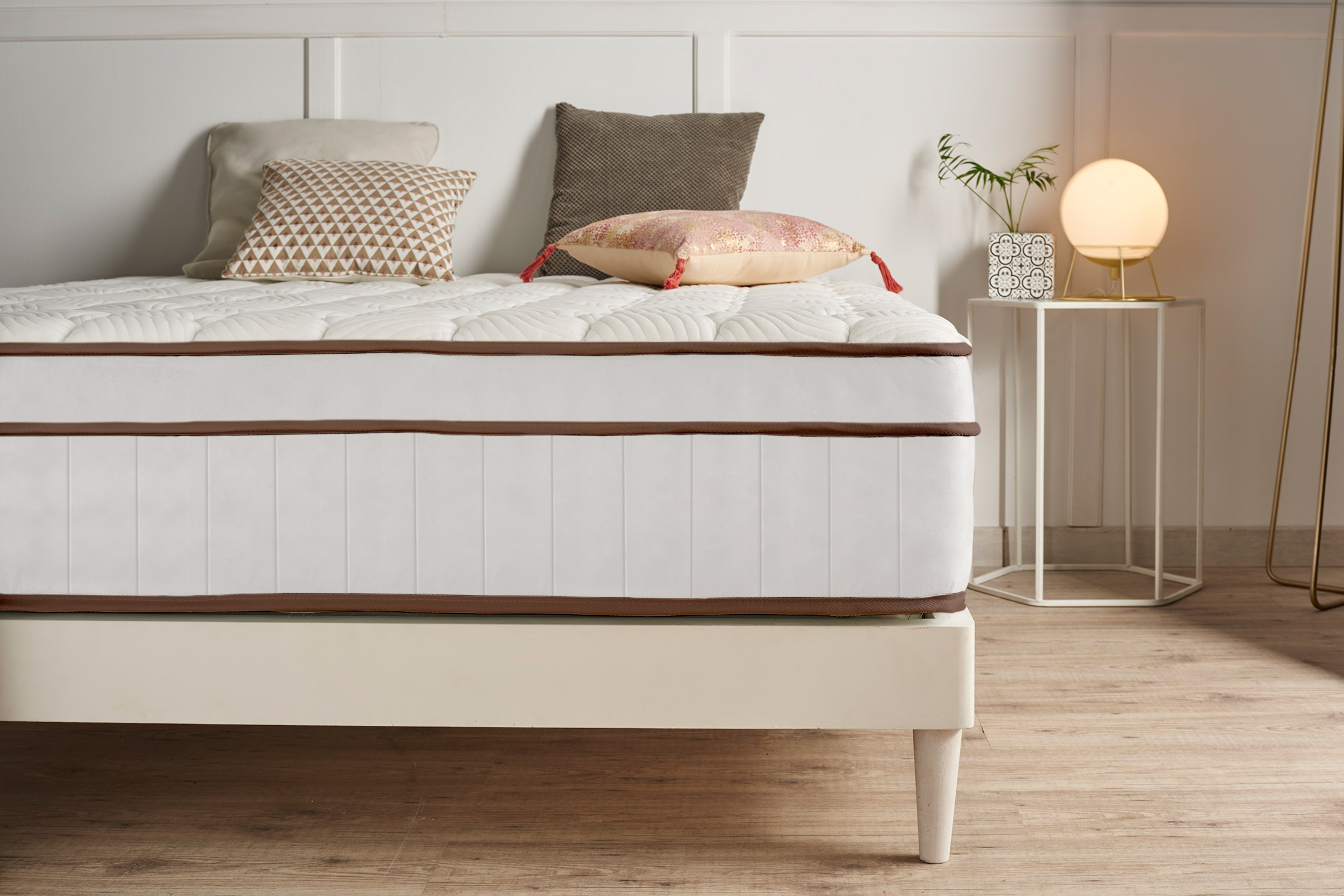 Un colchón blanco grueso con ribete marrón sobre una cama de madera, decorado con cojines y una lámpara.