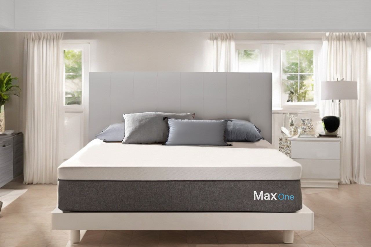 Una cama Max One con base gris, colchón blanco y cojines en un dormitorio luminoso con cortinas blancas.
