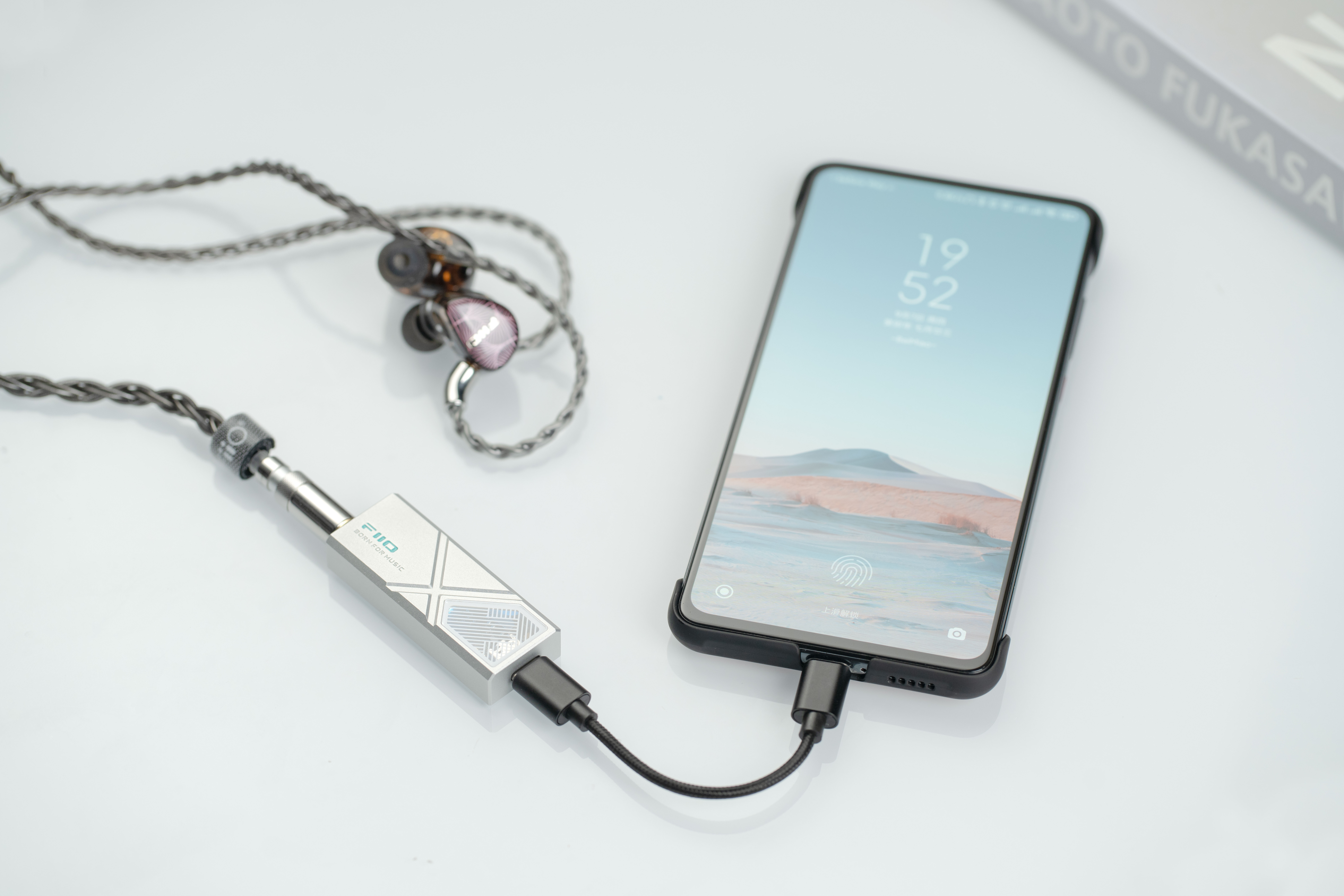 Smartfon podłączony do srebrnego przenośnego wzmacniacza DAC ze słuchawkami na białej powierzchni.