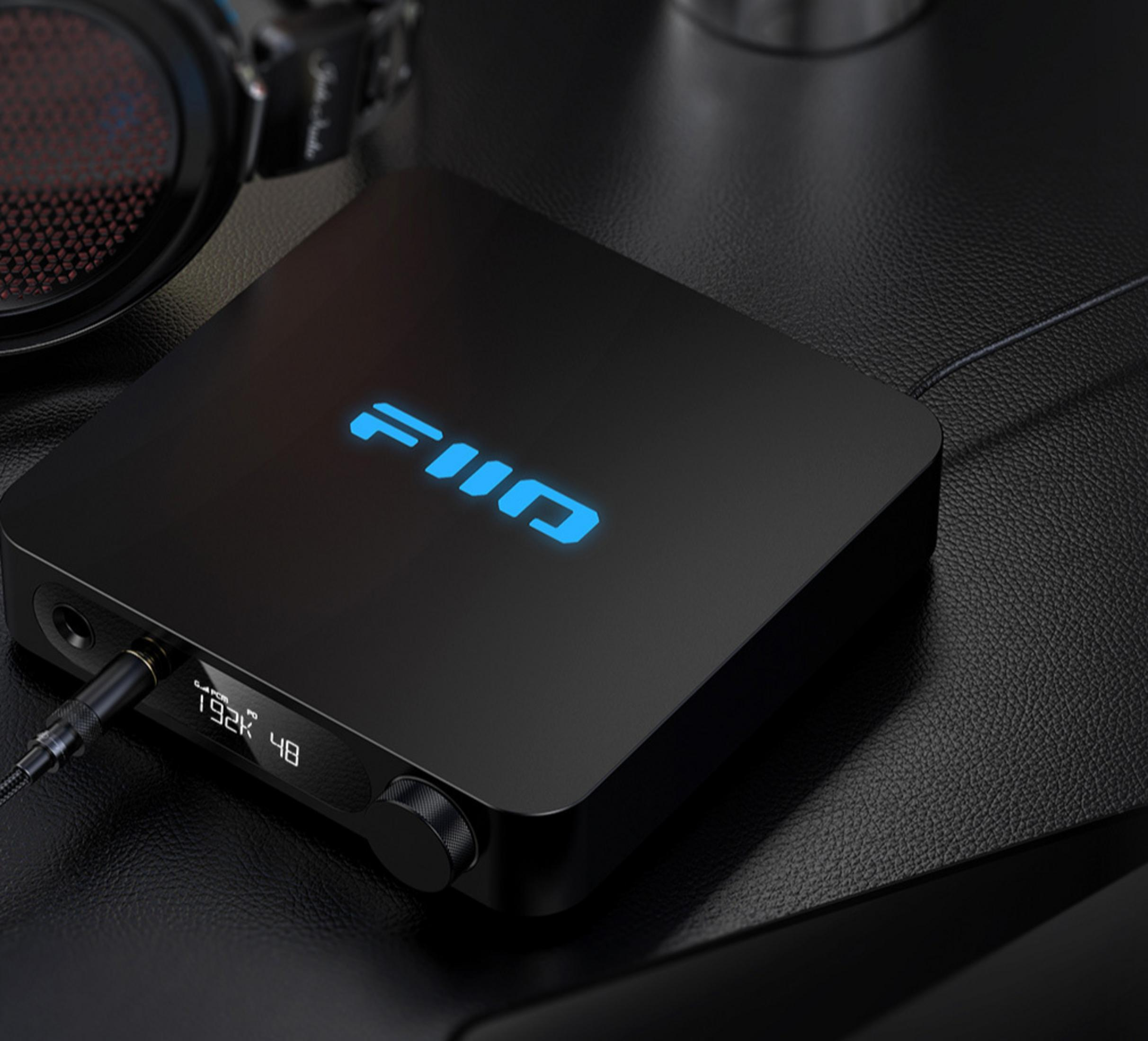 Czarny wzmacniacz audio FiiO z świecącym niebieskim logo na skórzanym biurku, podłączony do słuchawek.