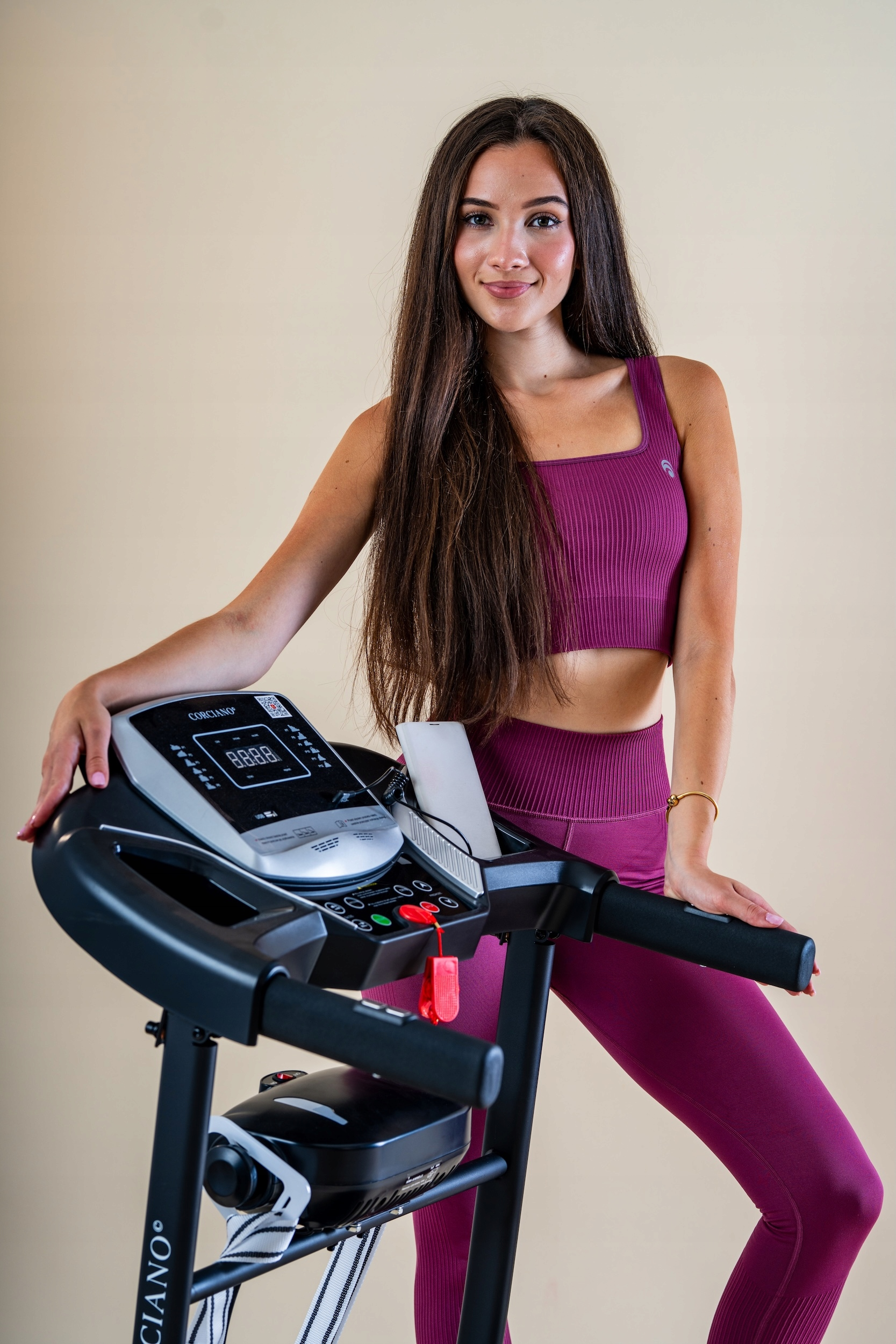 Uśmiechnięta brunetka w różowym stroju sportowym pozuje przy bieżni na beżowym tle.