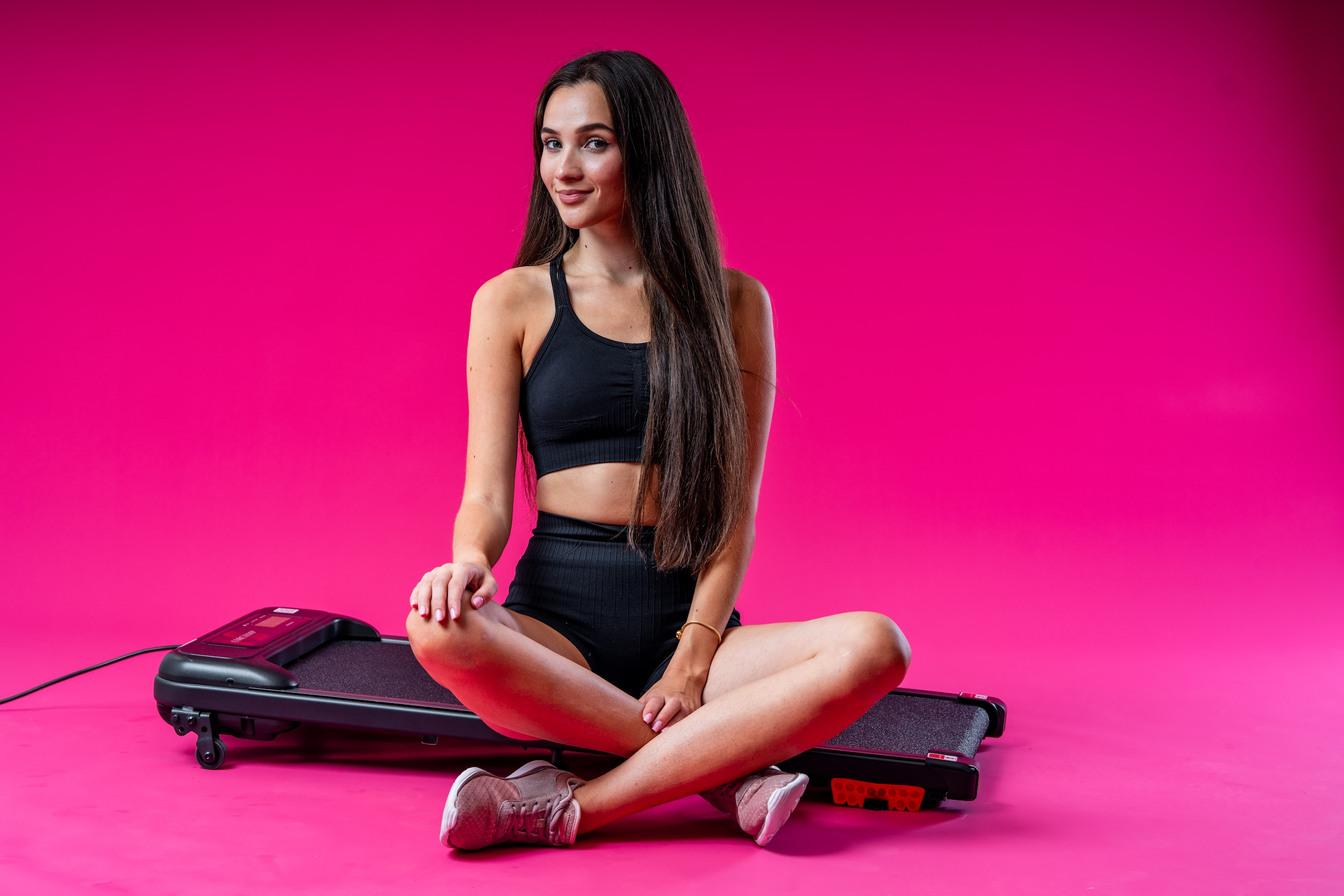 Brunetka w czarnym stroju sportowym siedzi na bieżni na różowym tle.