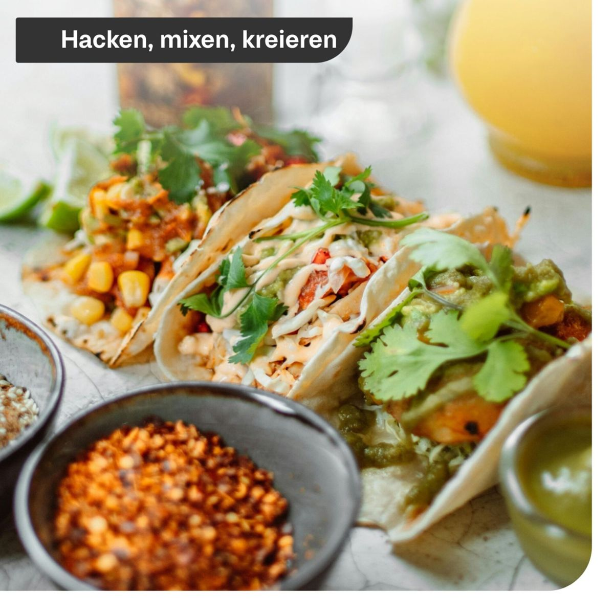 Drei gefüllte Tacos auf einem Tisch mit kleinen Schalen voller Gewürze und Kräutern garniert.