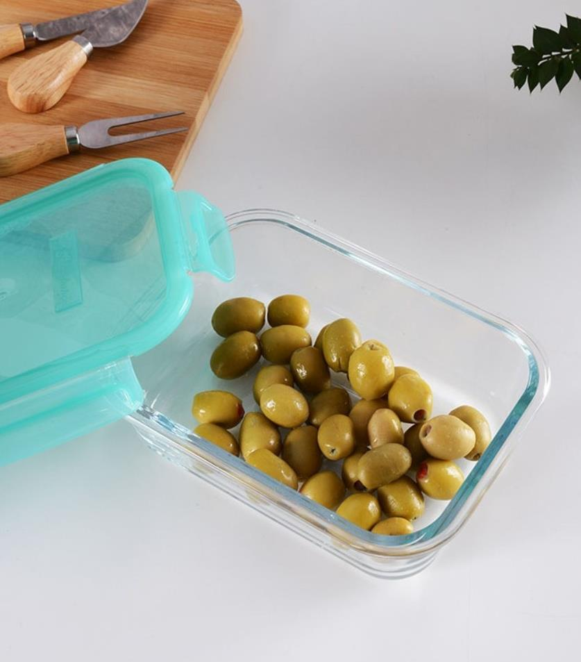 Açık renkli masada turkuaz kapaklı açık cam kap içinde yeşil zeytin.