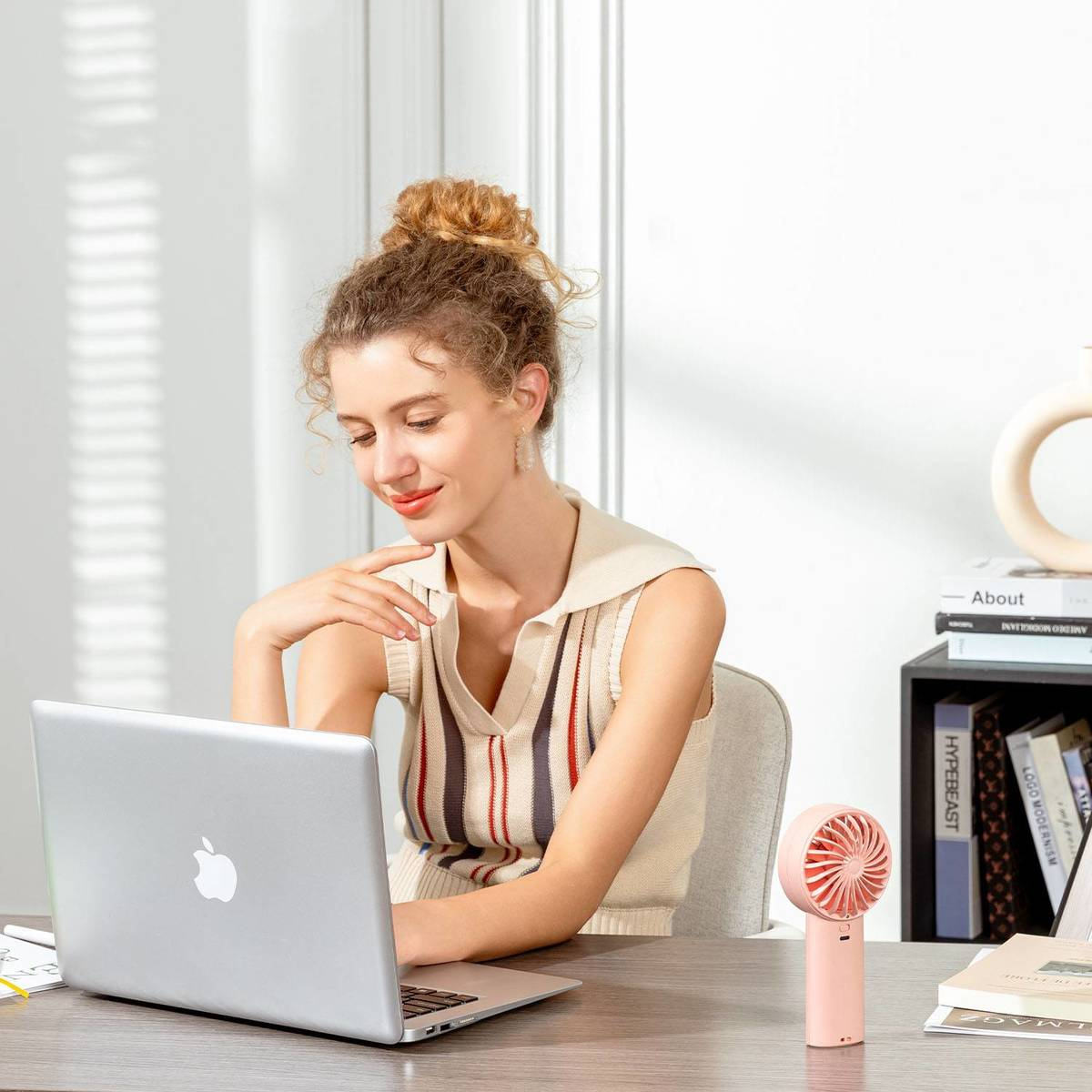 Eine blonde Frau arbeitet an einem Laptop an einem Holztisch, neben ihr steht ein kleiner rosa Ventilator.