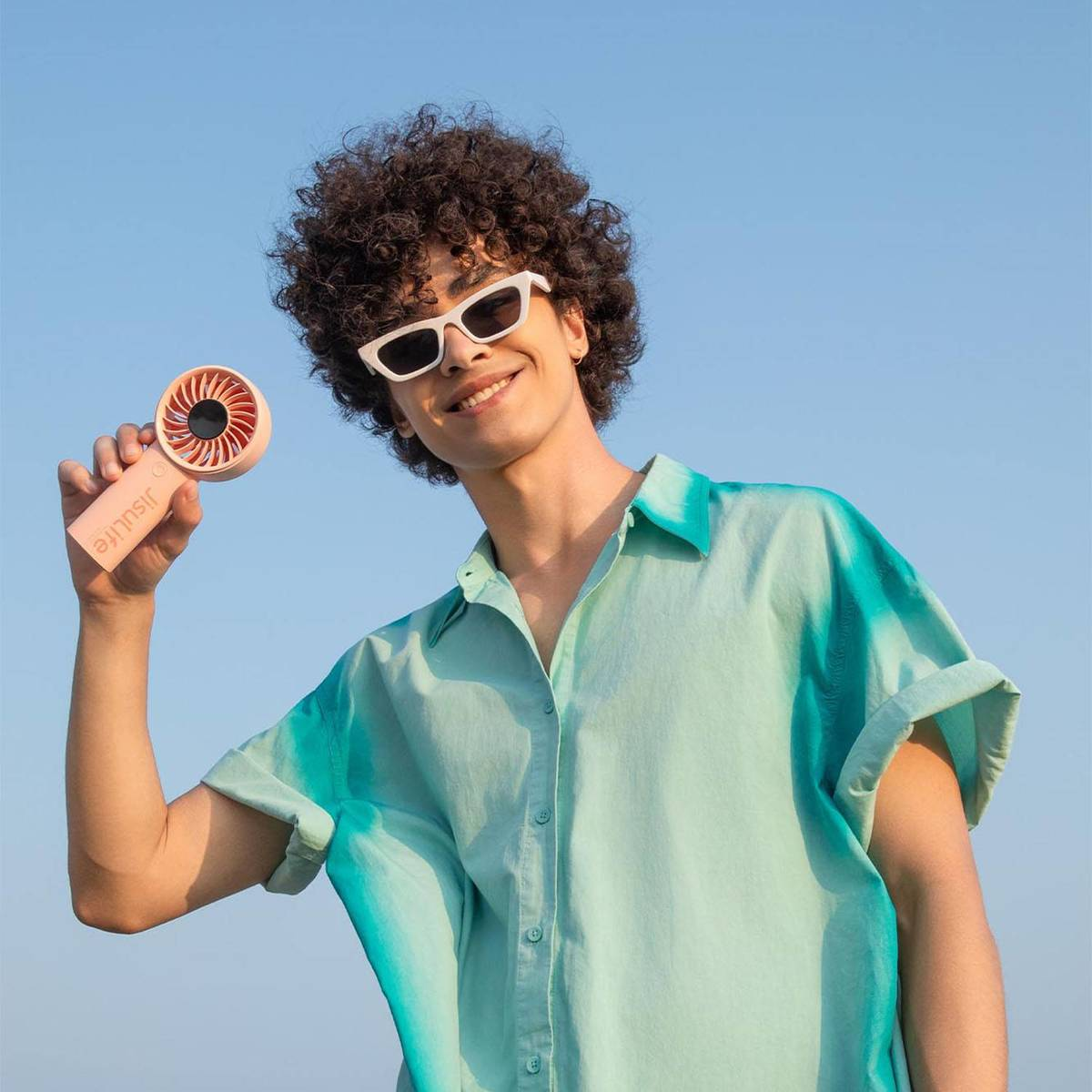 Ein junger Mann mit lockigem Haar und Sonnenbrille lächelt und hält einen kleinen rosafarbenen Handventilator.