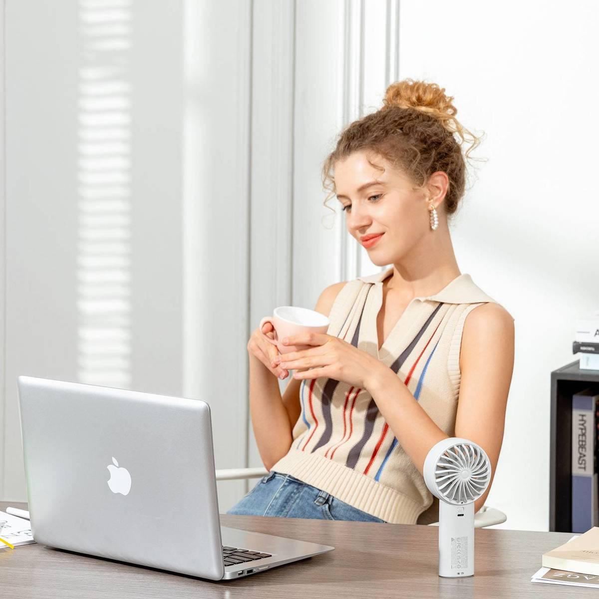 Eine Frau sitzt am Schreibtisch, trinkt aus einer Tasse und schaut auf einen Laptop neben einem kleinen Ventilator.