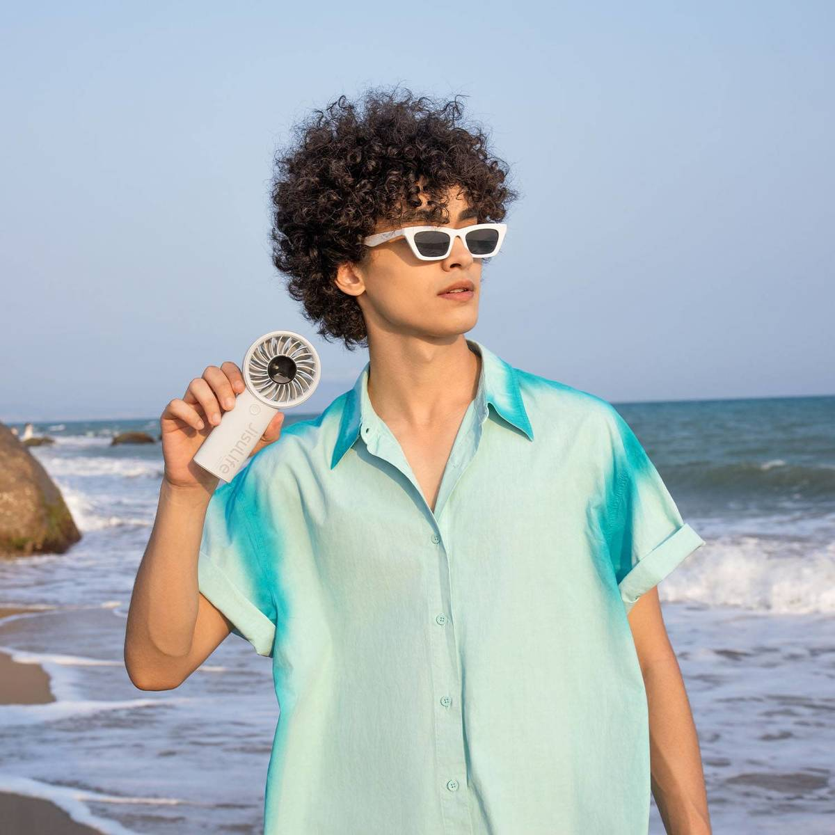Ein lockiger junger Mann mit weißer Sonnenbrille und türkisem Hemd hält einen Handventilator am Meer.