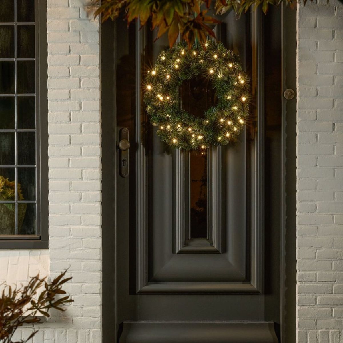 A lit Christmas wreath hangs on a dark door against a white brick wall.