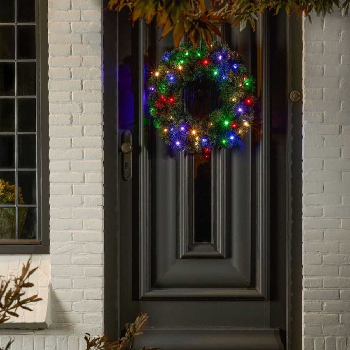 A lit Christmas wreath with colorful lights hangs on a dark front door.